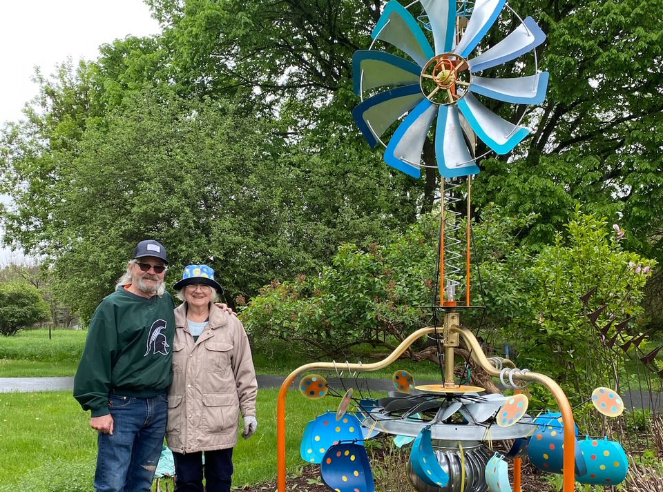 Man and woman standing in front of a windmill metal art piece in a flower garden